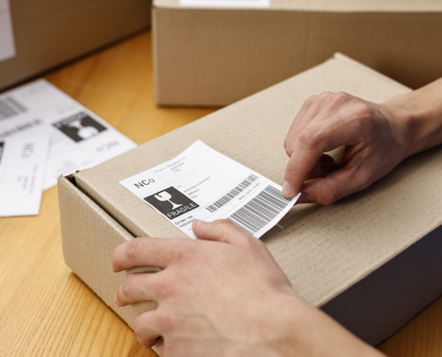 Man sticking shipping label with barcode on parcel at wooden table indoors, closeup